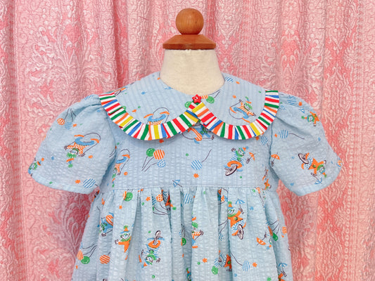 Children's babydoll style dress made of a seersucker blue cotton vintage fabric with birthday clowns printed all over, the collar is peter pan style with ruffles all around and a red star button detail at the center. Dress is on a mannequin against a pink patterned background, closeup of the collar.