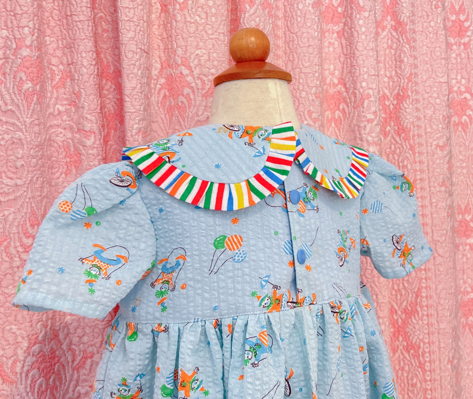 Children's babydoll style dress made of a seersucker blue cotton vintage fabric with birthday clowns printed all over, the collar is peter pan style with ruffles all around and a red star button detail at the center. Dress is on a mannequin against a pink patterned background, closeup of back details that includes blue snap closures.
