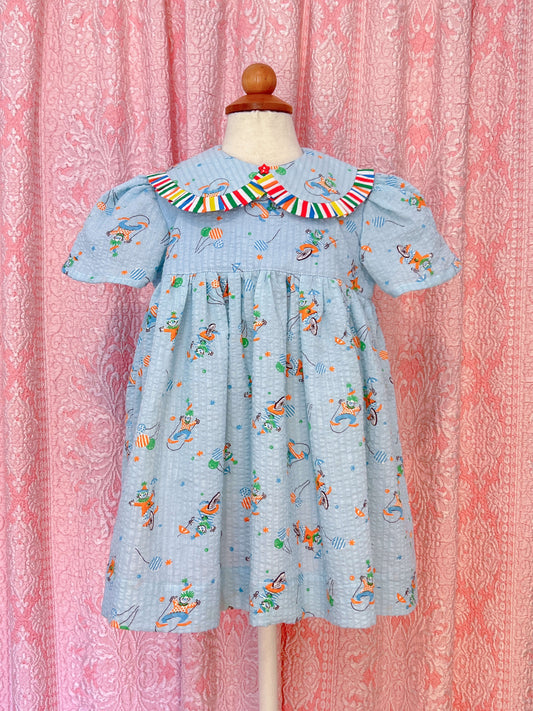 Children's babydoll style dress made of a seersucker blue cotton vintage fabric with birthday clowns printed all over, the collar is peter pan style with ruffles all around and a red star button detail at the center. Dress is on a mannequin against a pink patterned background